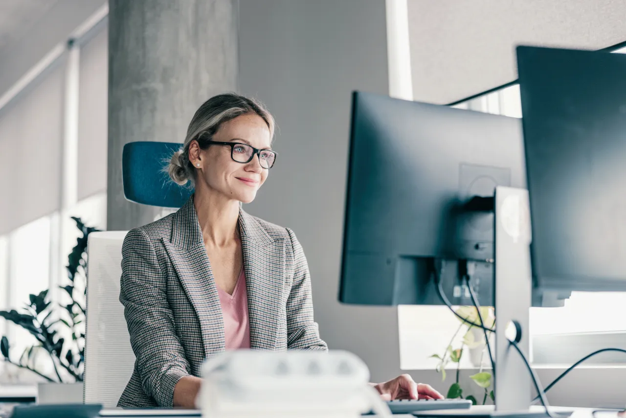 Uśmiechnięta kobieta w okularach, pracująca przy komputerze. Jej biurko jest uporządkowane, a w tle widać rośliny. To idealne miejsce dla księgowego.