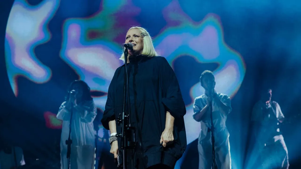 Kto śpiewa sprzedawcy marzeń? Wokalistka w czarnej, obszernej kreacji na tle psychodelicznej scenografii, z ch&oacute;rzystami w białych strojach.