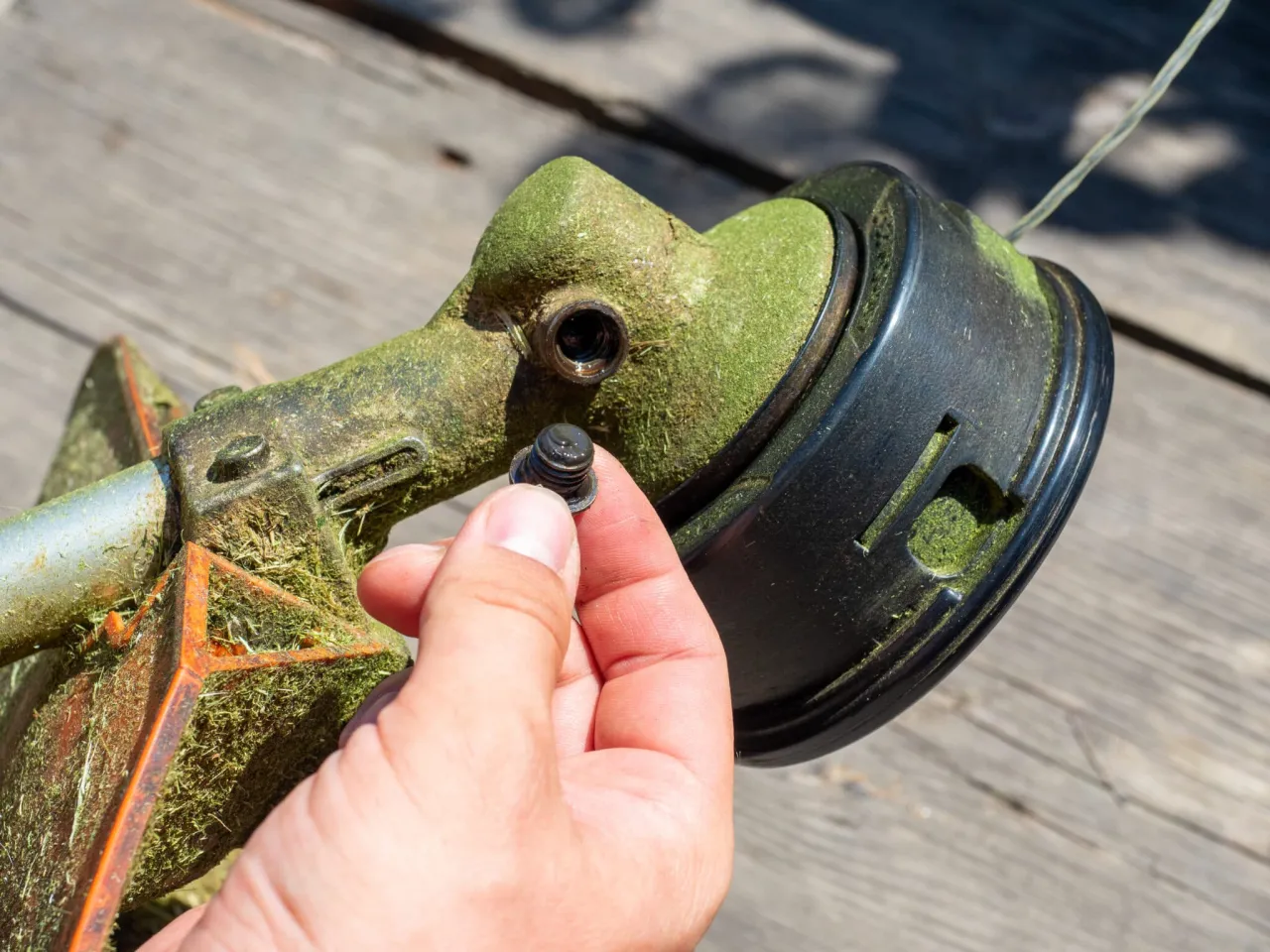 Dłoń trzyma śrubę, pokazując, jak odkręcić nóż od kosiarki. Widoczna głowica podkaszarki z trawą.