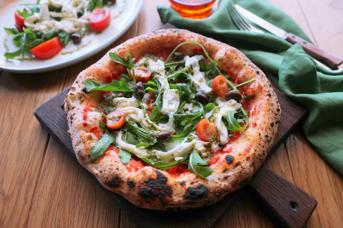 Pizza, gdzie powstała, z rukolą, pomidorkami i mozzarellą, na drewnianej desce.