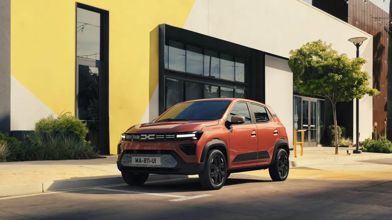 Pomarańczowy, elektryczny Dacia Spring, kt&oacute;ry może być odpowiedzią na pytanie, ile kosztuje najtańszy samoch&oacute;d elektryczny, zaparkowany przed nowoczesnym budynkiem.