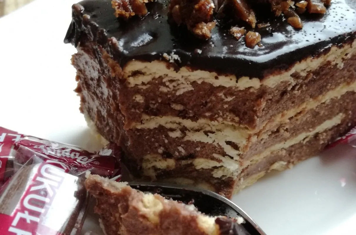 Czekoladowe ciasto kukułka z kremem i wafelkami, polane lukrem, z okruszkami na wierzchu. Obok opakowanie cukierk&oacute;w Kukułka.