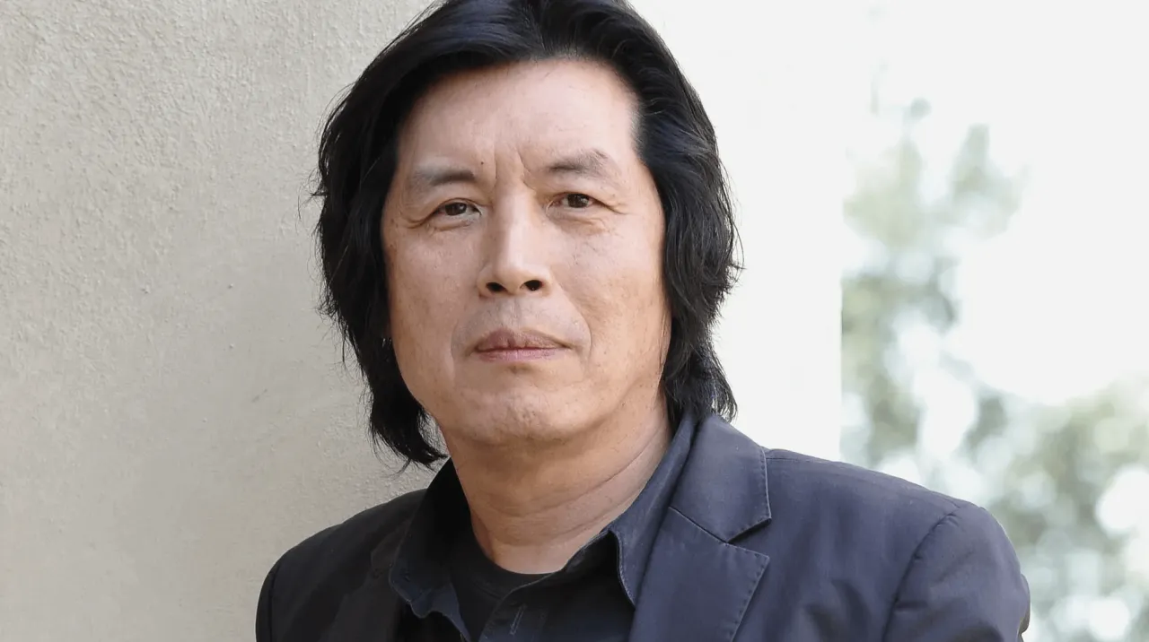 Portret reżysera Lee Chang Dong, zamyślony, w ciemnej marynarce, na tle jasnej ściany i rozmytej zieleni.