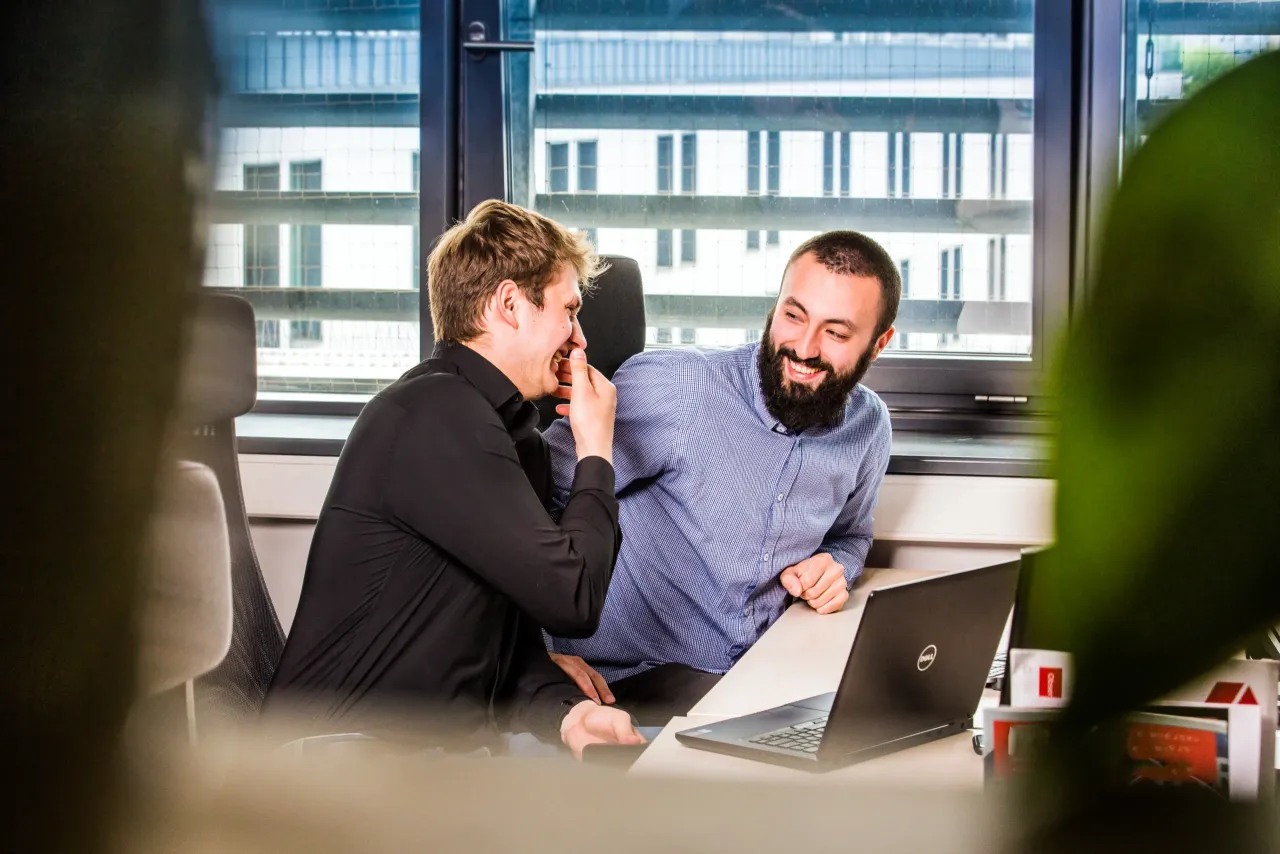 Dw&oacute;ch mężczyzn śmieje się podczas szkolenia GA4. Jeden z nich wskazuje na ekran laptopa, drugi opiera się o biurko.