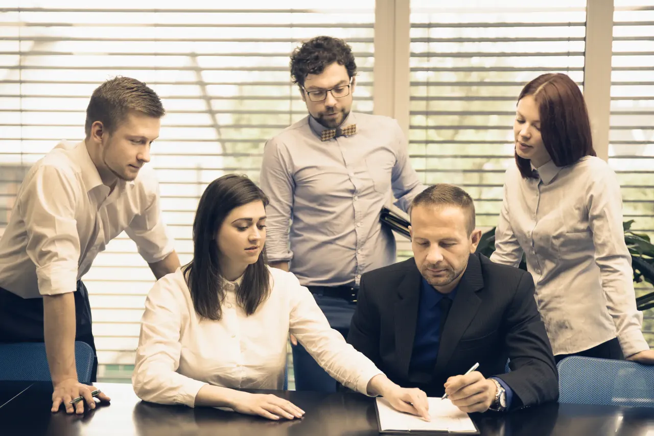 Zespół ludzi przy stole, mężczyzna podpisuje dokumenty. Sytuacja przypomina nabycie spadku u notariusza.