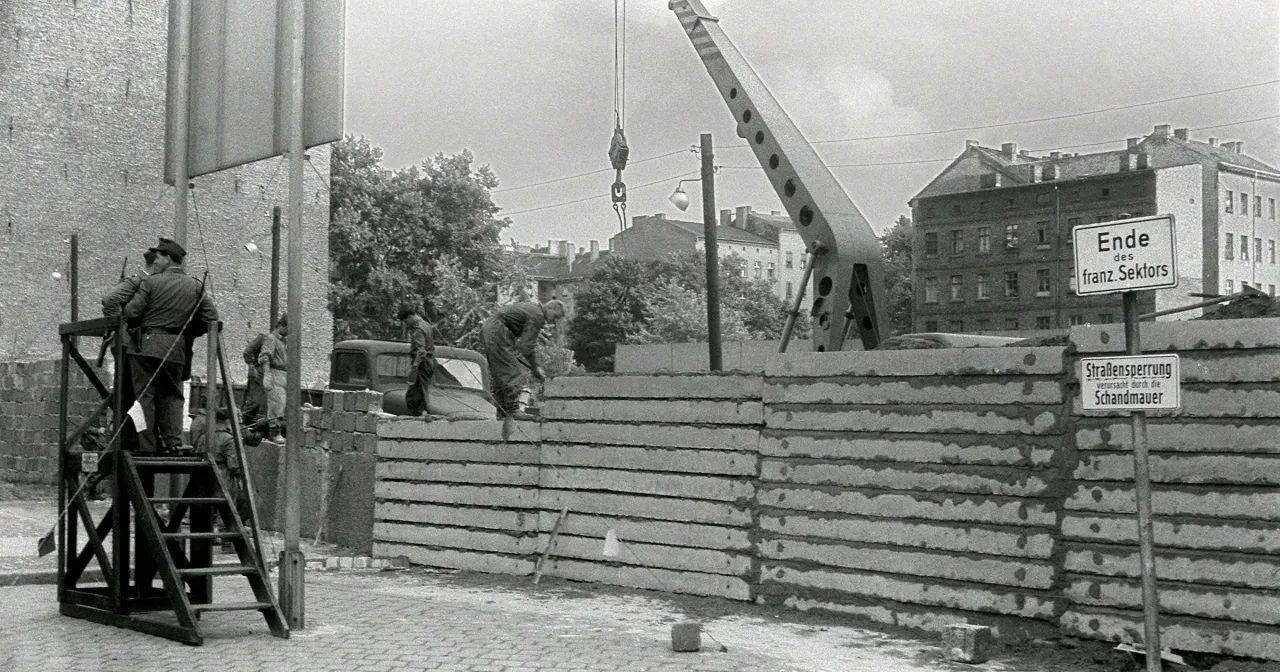 Budowa Muru Berlińskiego, widok na plac budowy z żołnierzami na wieżyczce i pracownikami układającymi betonowe płyty.