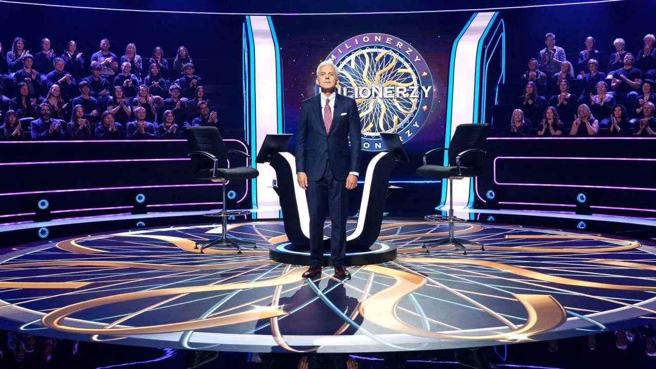 Hubert Urbański na scenie Milioner&oacute;w. Widownia w tle. Koła ratunkowe w Milionerach to: 50:50, telefon do przyjaciela, pomoc publiczności.