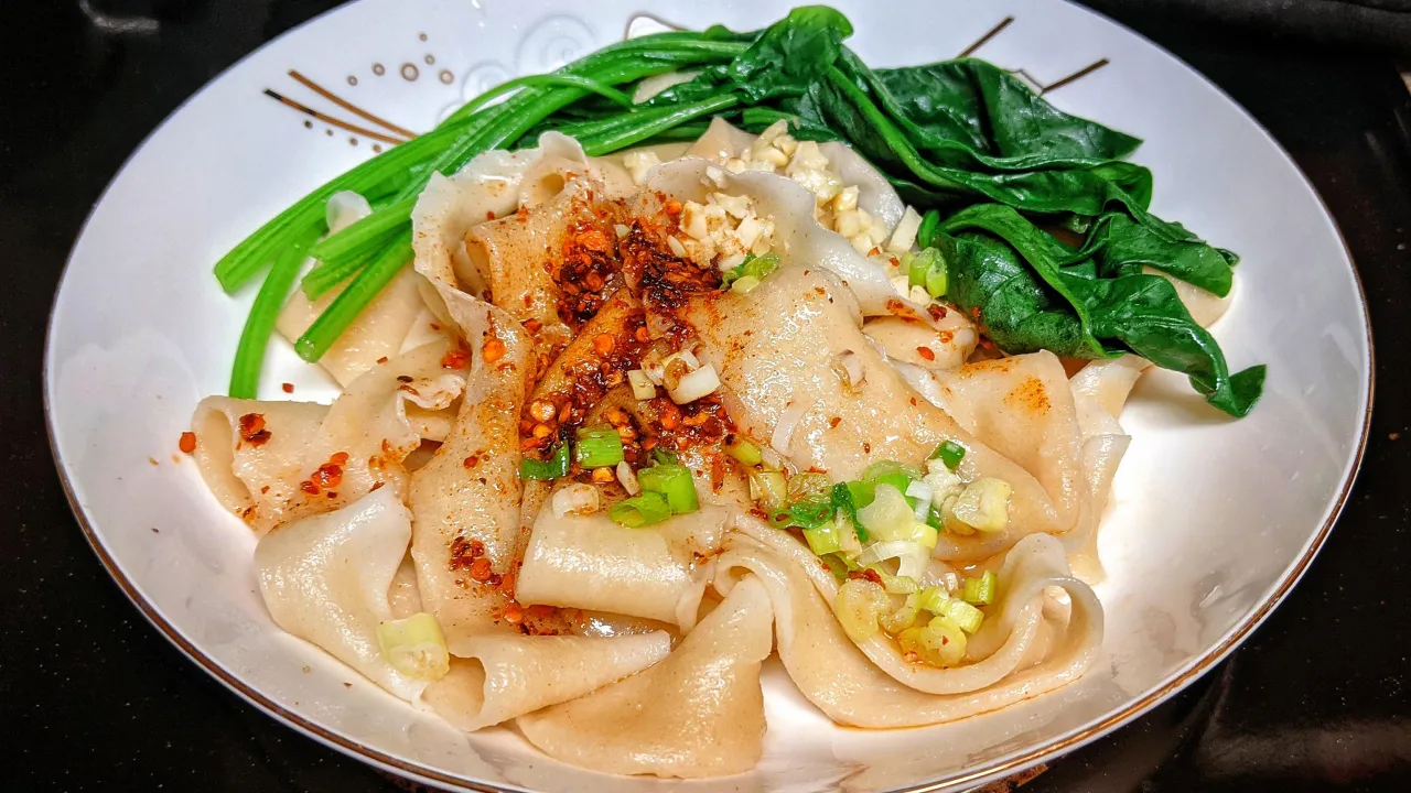 Szerokie, ręcznie robione kluski biang biang z czosnkiem, chilli i zieloną cebulką, podane z gotowanym szpinakiem.