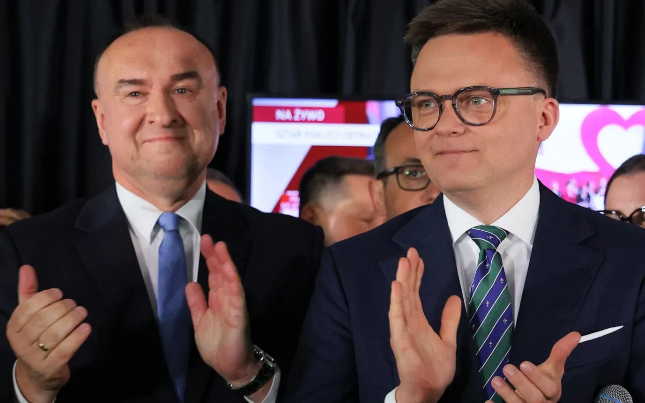 Dwóch mężczyzn w garniturach klaszcze. W tle ekran z napisem 