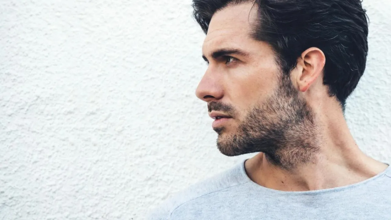 Mężczyzna z 3-dniowym zarostem, który ma około 2-3 mm długości, na tle białej ściany.