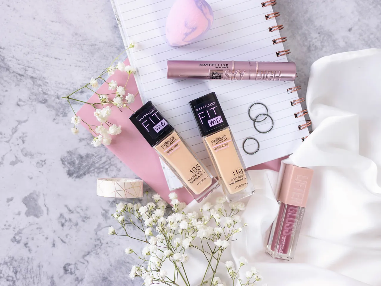 Maybelline Fit Me podkład rozświetlający, tusz do rzęs Sky High i błyszczyk Lifter Gloss na tle notatnika i kwiat&oacute;w.