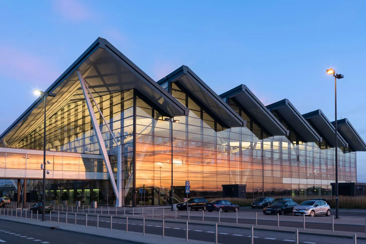 Nowoczesny budynek lotniska w Gdańsku z przeszkloną fasadą odbijającą zachodzące słońce.