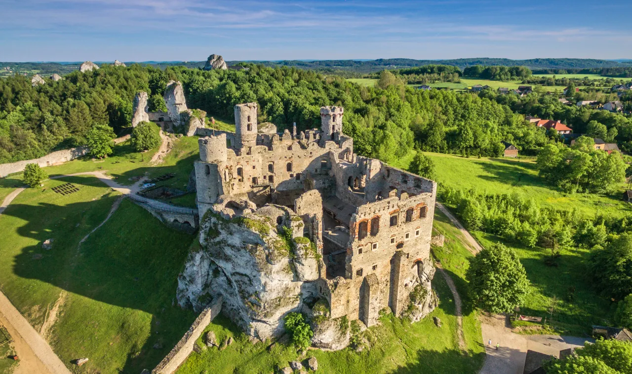 Ruiny zamku na skale, część trasy rowerowej Szlak Orlich Gniazd, otoczone zielonymi lasami i polami.