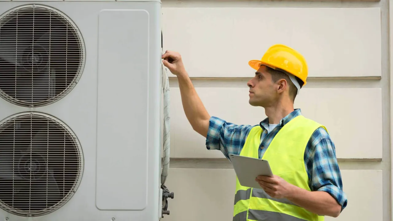 Technik w żółtym kasku i kamizelce odblaskowej sprawdza pompę ciepła. Regularny przegląd pompy ciepła zapewnia jej sprawność.