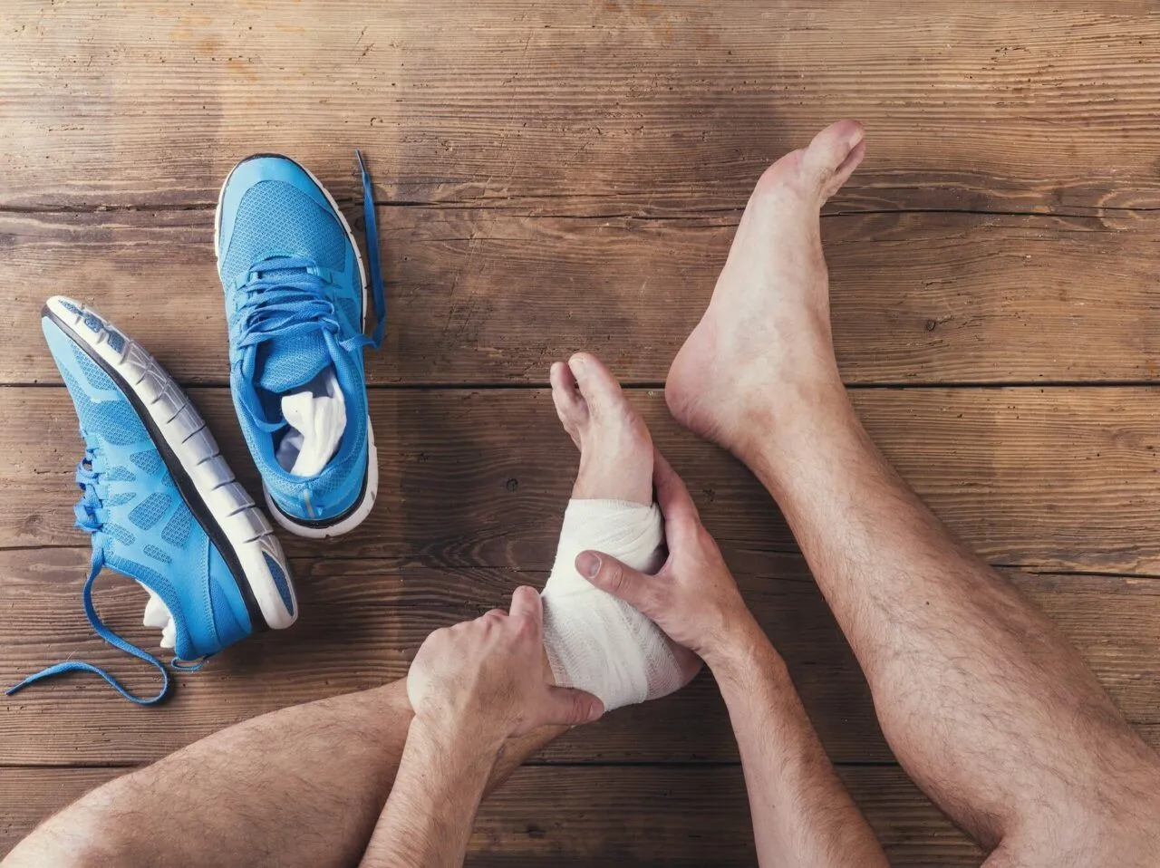 Skręcona kostka z opuchlizną, owinięta bandażem. Obok leżą niebieskie buty sportowe.