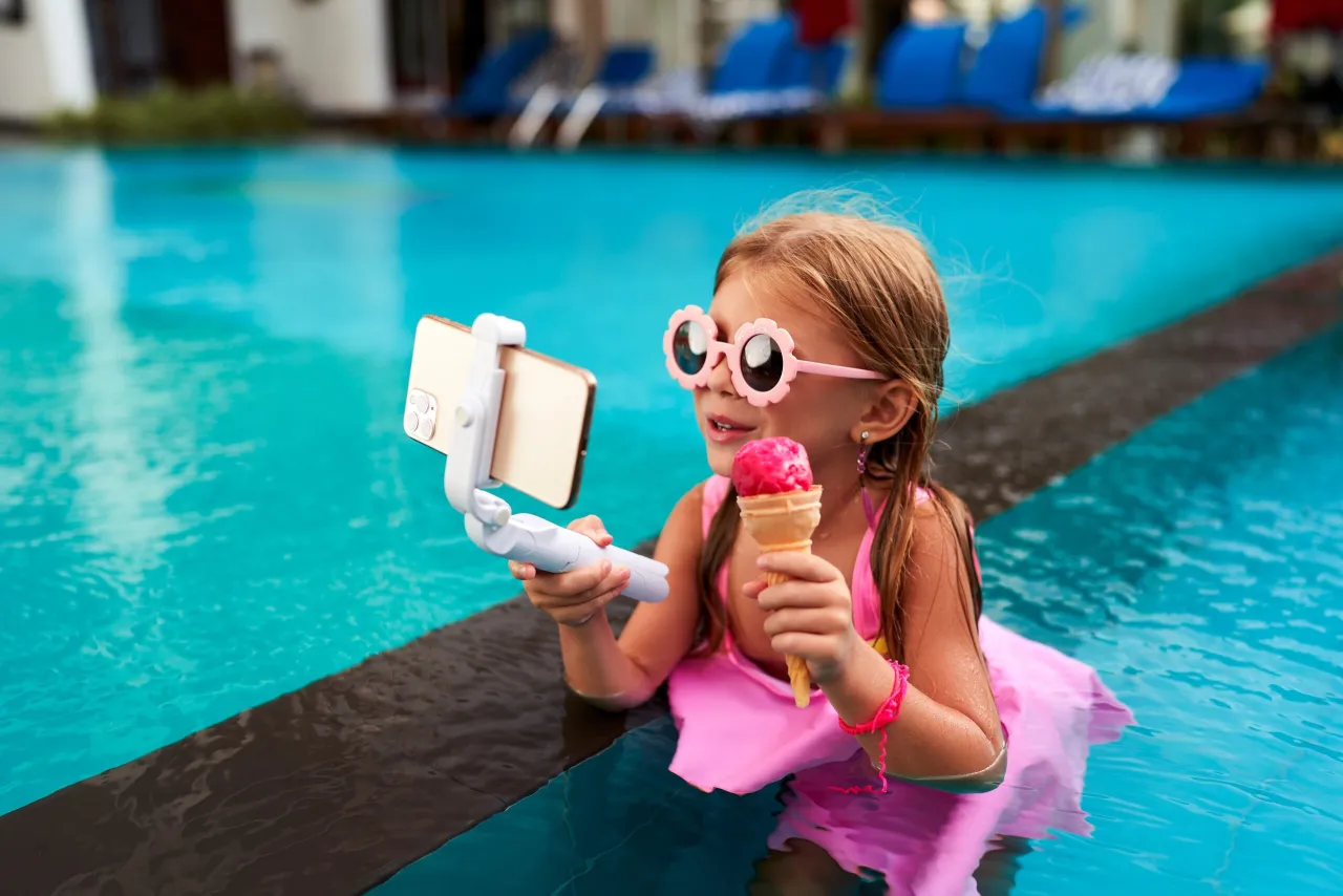 Mała influencerka w okularach w kształcie kwiatków nagrywa filmik z lodami w basenie, pokazując, ile zarabiają dzieci influencerów za kampanie.