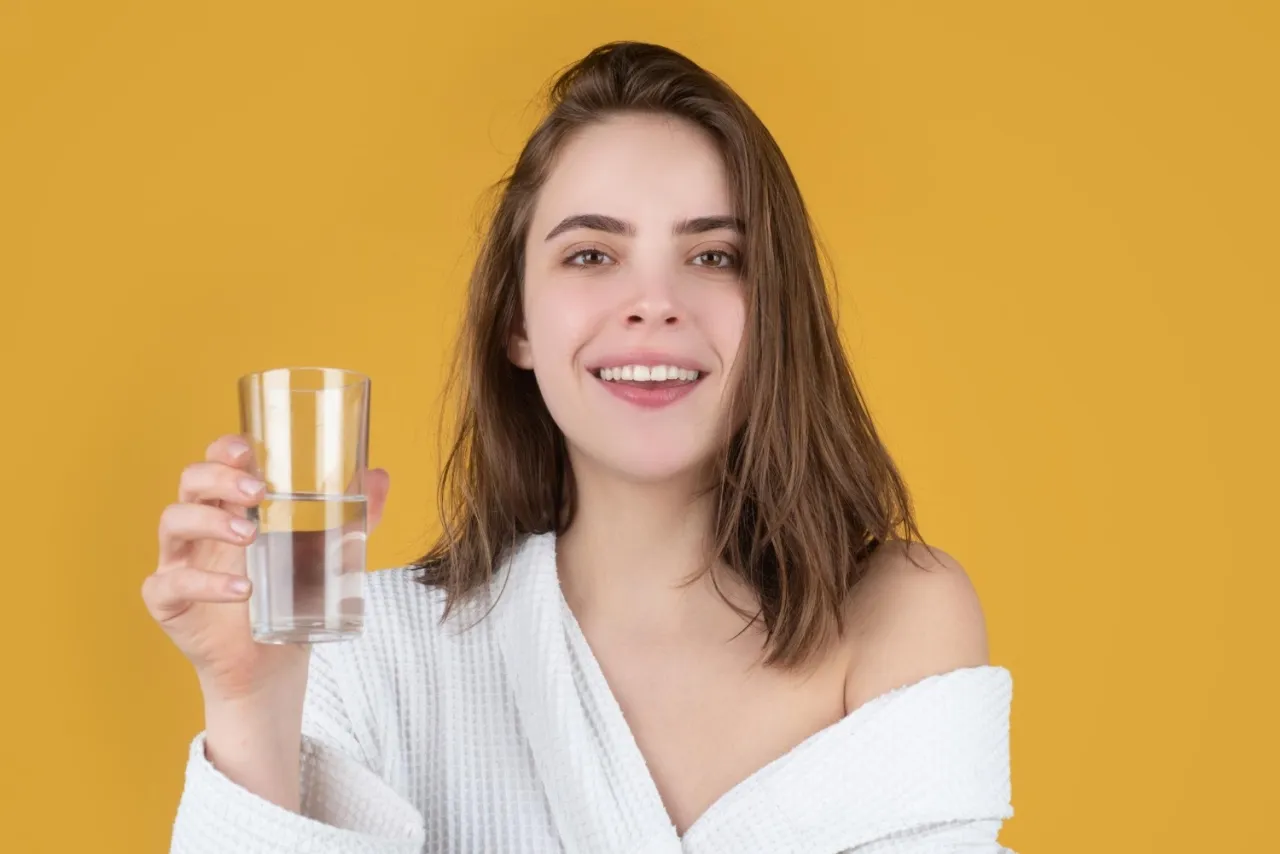 Uśmiechnięta kobieta w szlafroku trzyma szklankę wody, przypominając, kiedy pić kolagen dla zdrowej sk&oacute;ry.