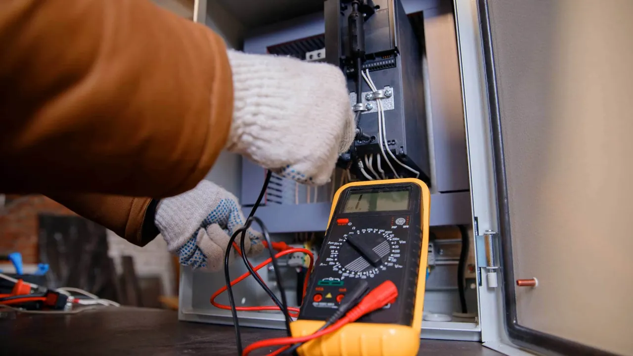 Ręce w rękawiczkach podłączają przewody do panelu sterowania, obok leży żółty multimetr. Pokazuje, jak używać multimetru do pomiarów.