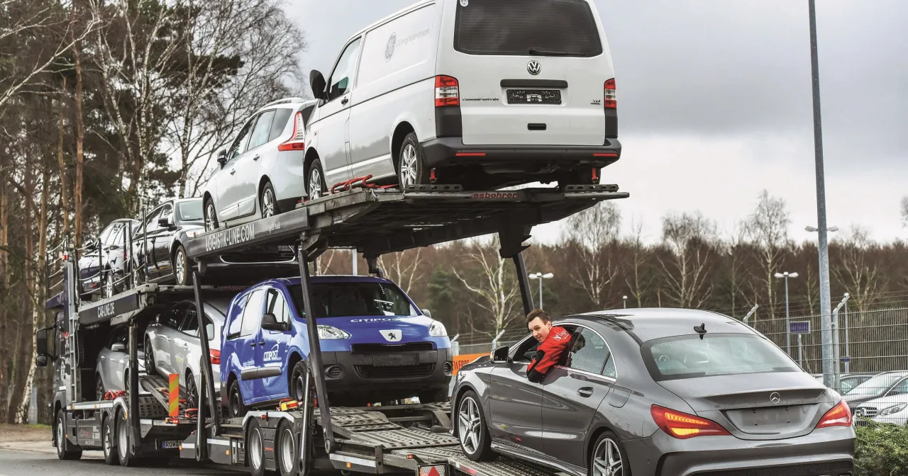 Sprowadzenie auta z Niemiec: laweta z samochodami, w tym Volkswagen Transporter i Peugeot Partner. Kierowca w czerwonej kurtce pozdrawia.