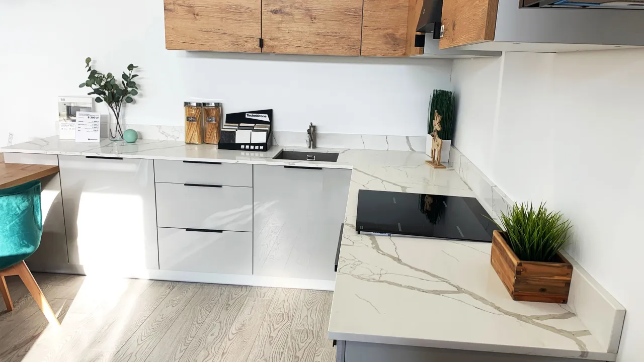 Nowoczesna kuchnia z białymi frontami i blatem z konglomeratu na podłogę. Widać zlew, płytę indukcyjną i drewniane szafki.