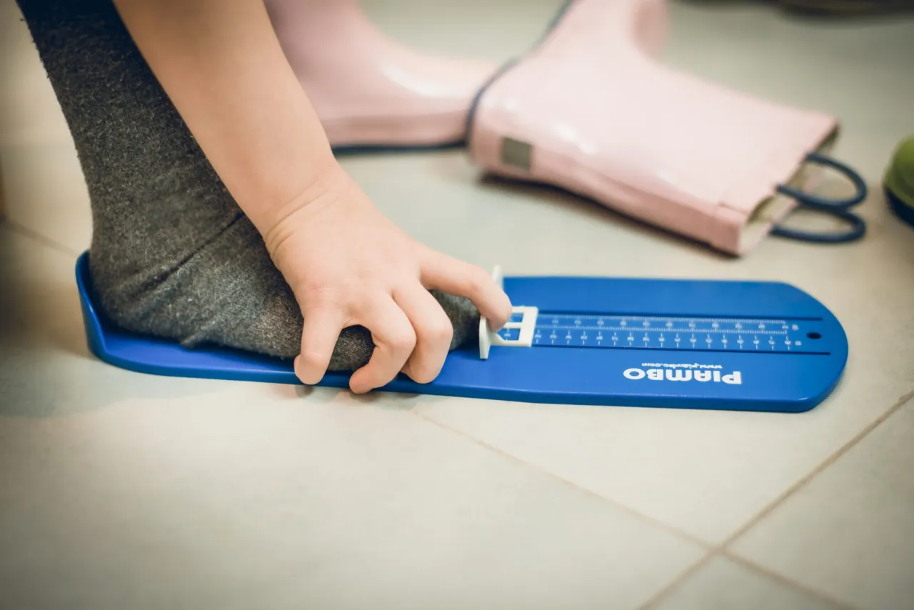 Dziecko mierzy stopę na niebieskim przyrządzie, aby sprawdzić, co to jest długość wkładki buta. W tle różowe kalosze.