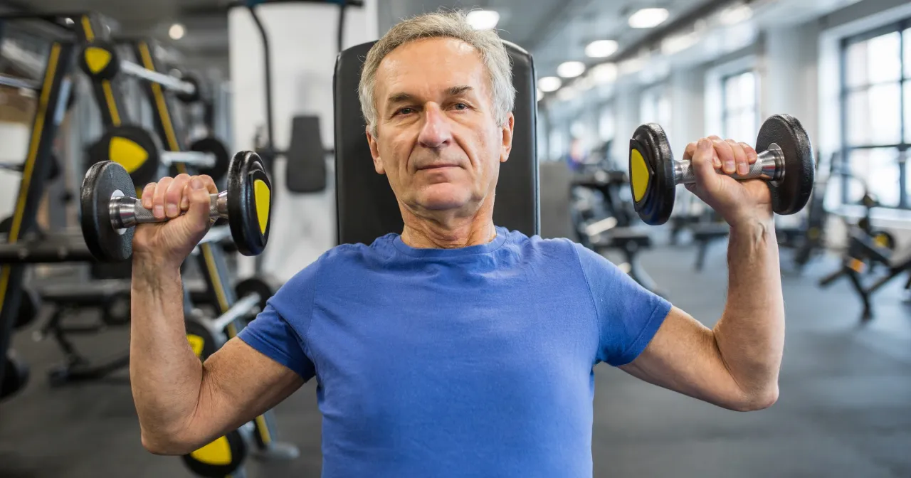 Starszy mężczyzna ćwiczy z hantlami na siłowni. Zastanawiasz się, kreatyna ile przed treningiem będzie najlepsza?
