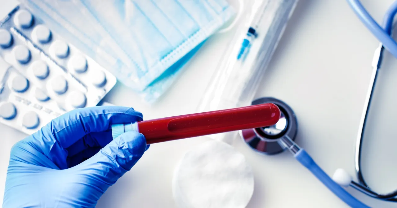 Rękawiczka z niebieskiej gumy trzyma prob&oacute;wkę z krwią. Obok leżą maseczka, tabletki, strzykawka i stetoskop. To może być część badania ast.