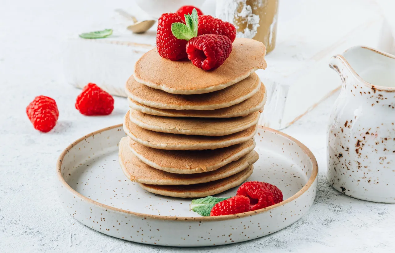 Stos puszystych placuszków, idealnych do przygotowania jak zrobic proteinowe pancakes, ozdobiony malinami i miętą. Obok dzbanuszek z syropem.