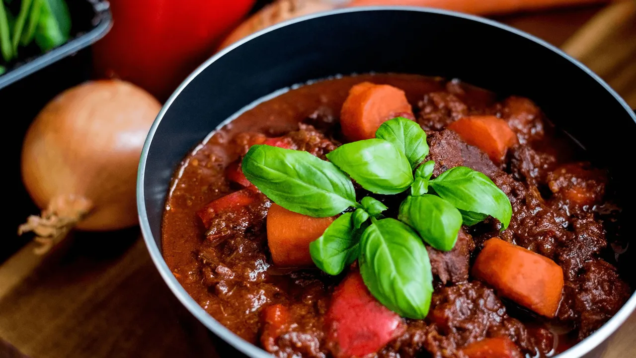 Gęsty, aromatyczny gulasz z wołowiny z kawałkami marchewki i papryki, udekorowany świeżą bazylią.