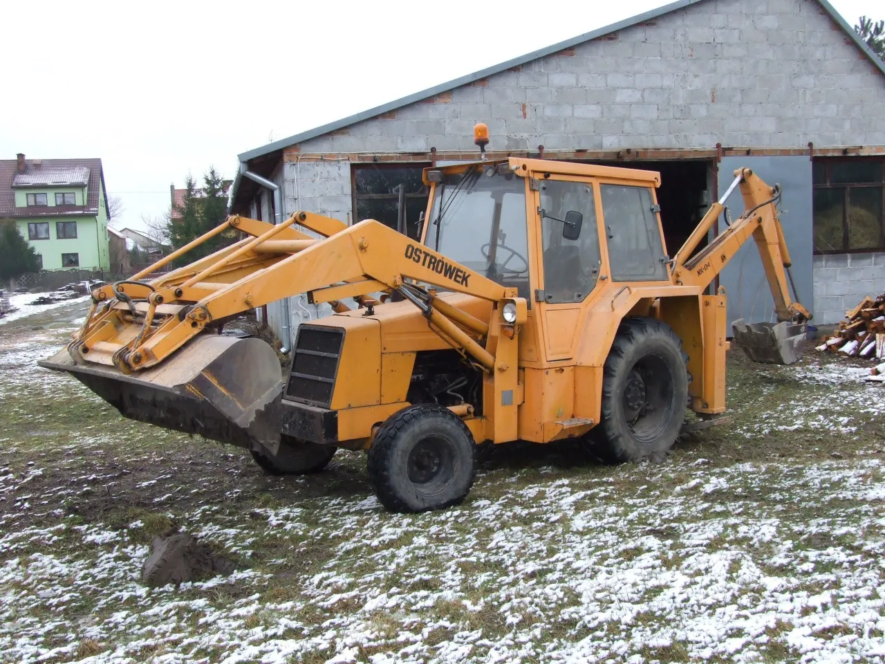 Żółta koparko-ładowarka Ostrówek stoi na zaśnieżonej trawie. Czy warto kupić koparkę Ostrówek? To solidna maszyna do prac ziemnych.