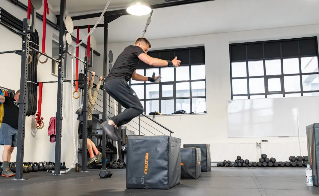 Mężczyzna wykonuje wyskok na skrzynię, ćwicząc w stylu cross trening co to jest. W tle widać sprzęt do ćwiczeń i innych trenujących.