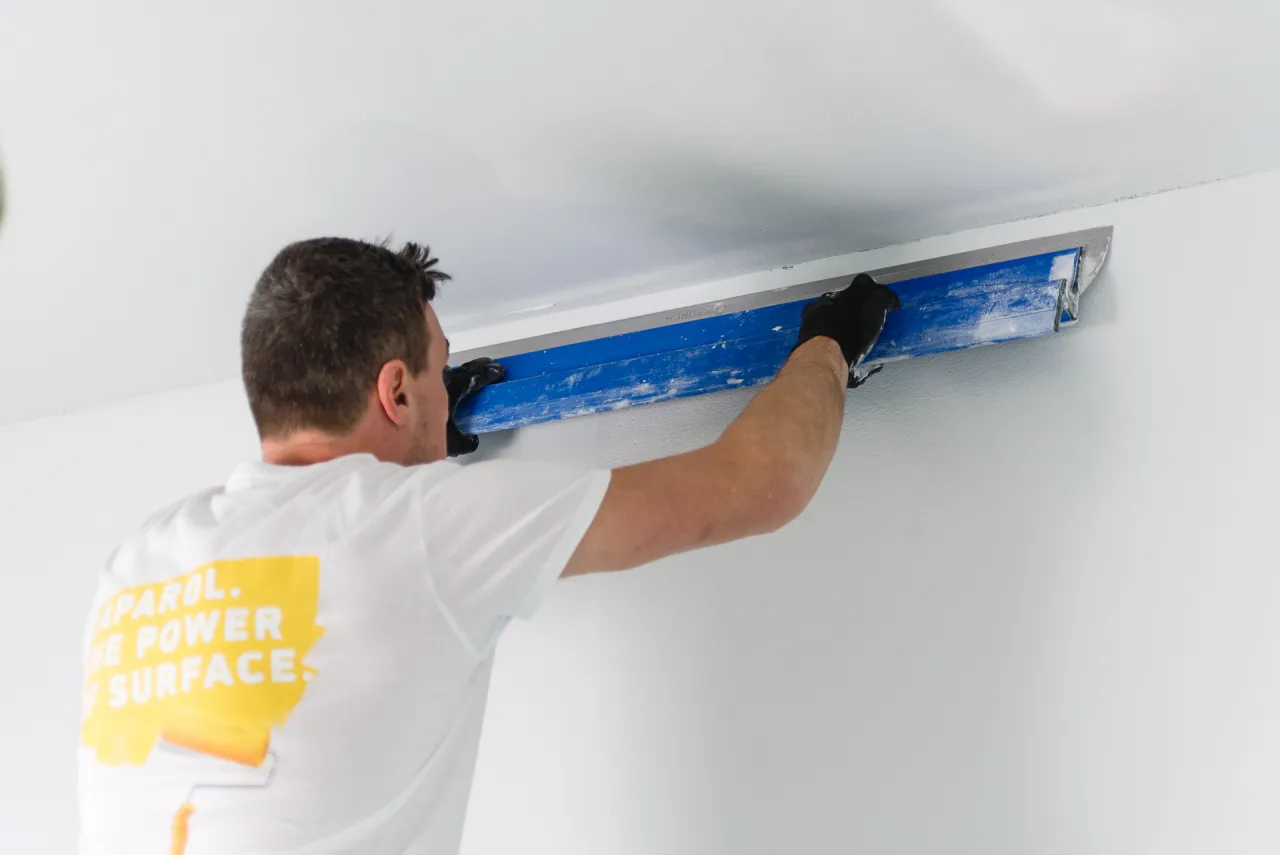 Mężczyzna w białej koszulce z logo Caparol wygładza sufitową listwę. Zastanawiasz się, jak często malować mieszkanie? To zależy od wielu czynników!