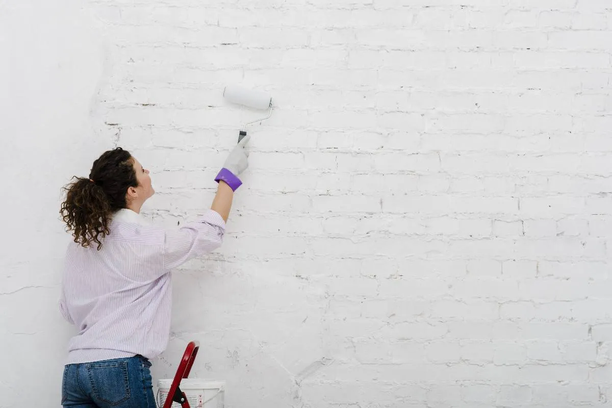 Kobieta maluje wałkiem białą ceglaną ścianę, pokazując, jak zmienić kolor kamienia dekoracyjnego gipsowego.