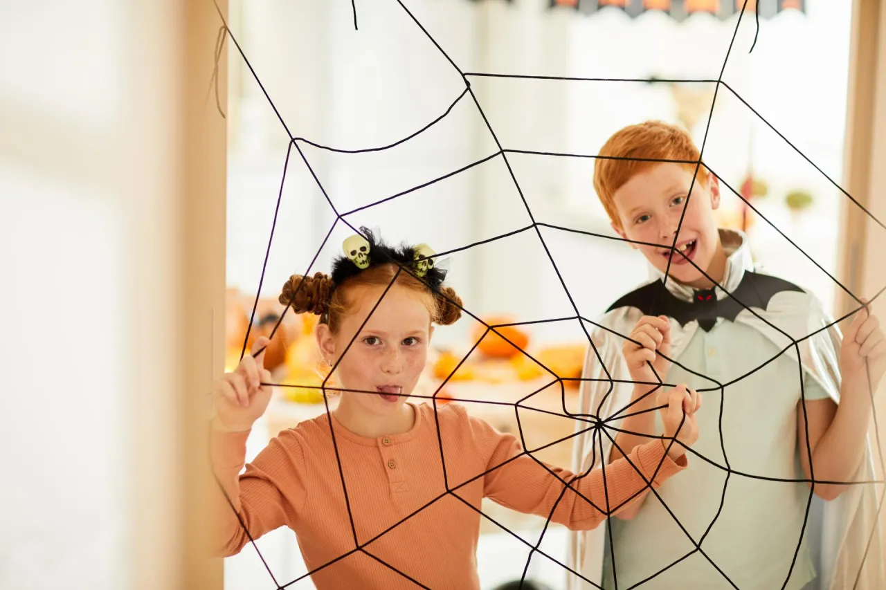 Dzieci w strojach na Halloween bawią się, trzymając się wielkiej pajęczyny. Dziewczynka z ozdobami w kształcie czaszek na głowie wyciąga język, a chłopiec w pelerynie wampira uśmiecha się szeroko.