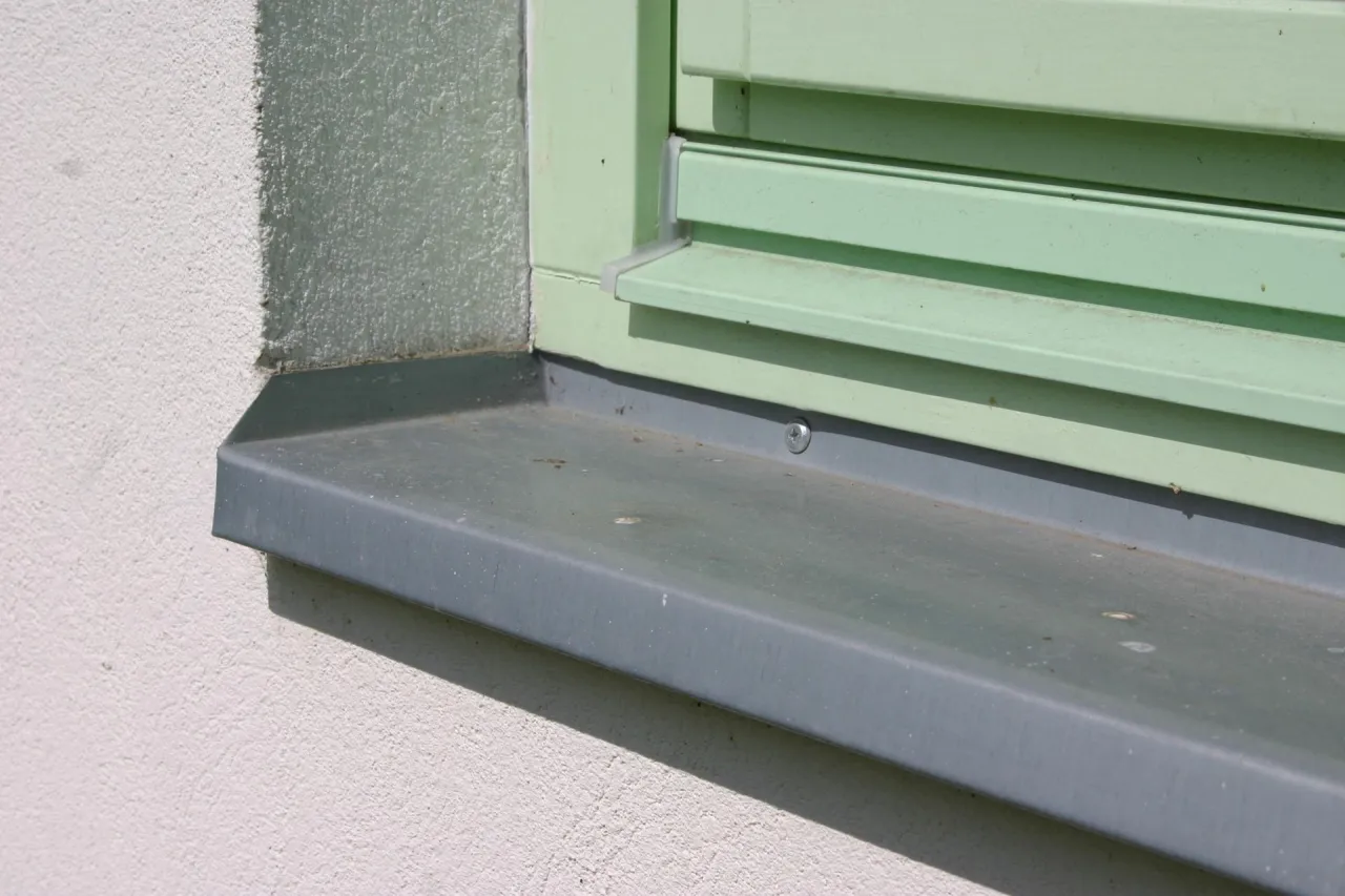 Szary parapet zewnętrzny zamontowany do czoła okna z zielonymi okiennicami.