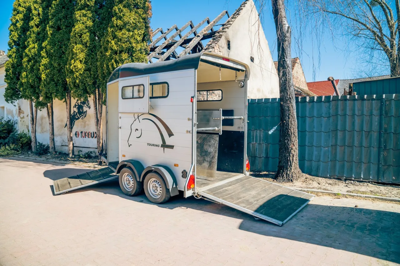 Otwarta przyczepa na konie z rampą, gotowa do załadunku. Obok drzewa i zniszczony budynek.