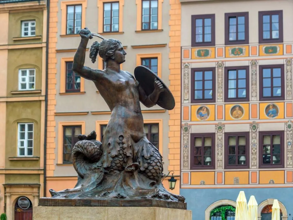 Pomnik Syrenki Warszawskiej, bohaterki legendy o Warsie i Sawie, z mieczem i tarczą, na tle kolorowych kamienic.