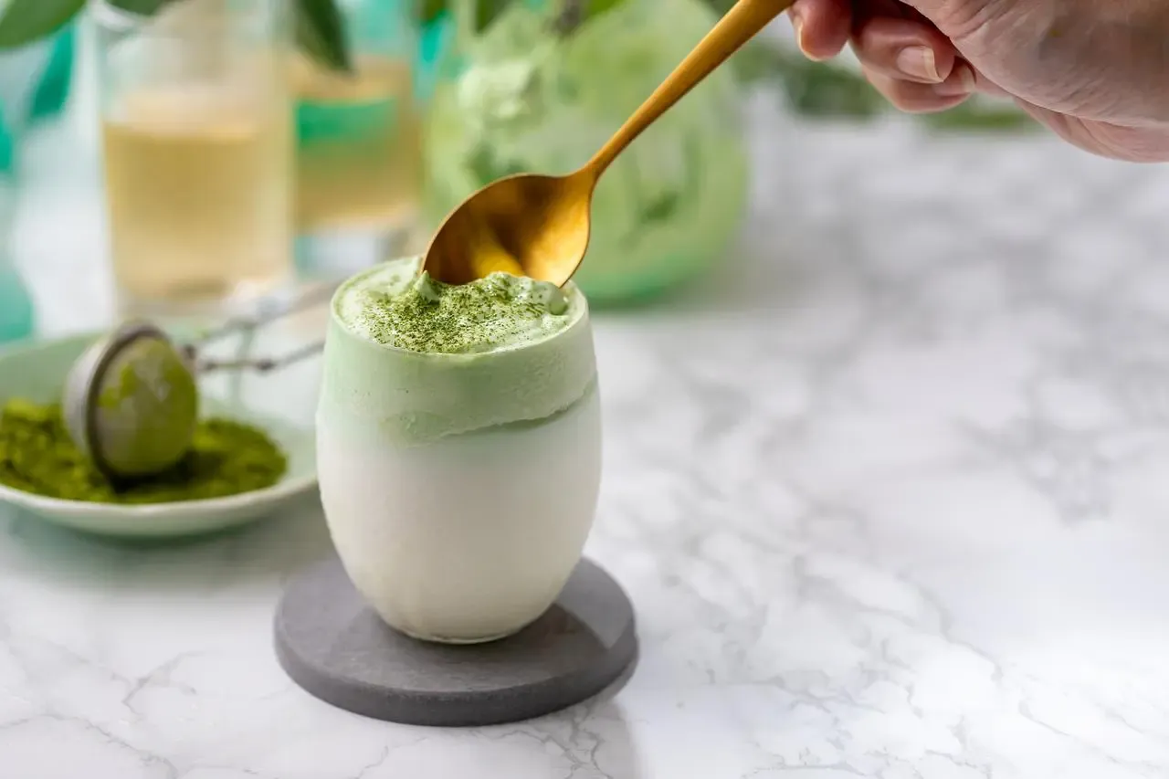Puszysty matcha latte, czyli jak zrobić ten popularny nap&oacute;j. Złota łyżka zanurza się w zielonej piance.