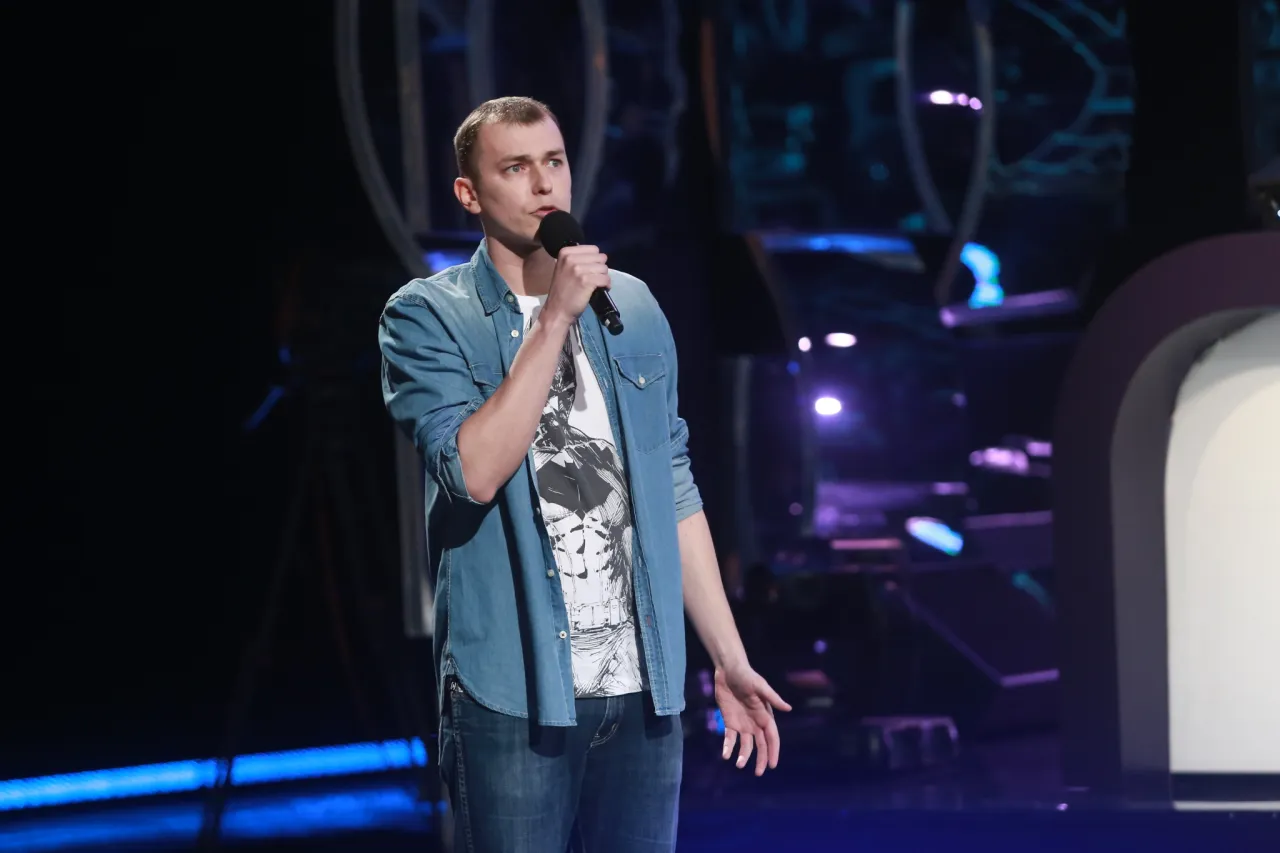 Rafał Banaś na scenie, w dżinsowej koszuli i z mikrofonem w dłoni, opowiada dowcipy podczas występu stand-up.
