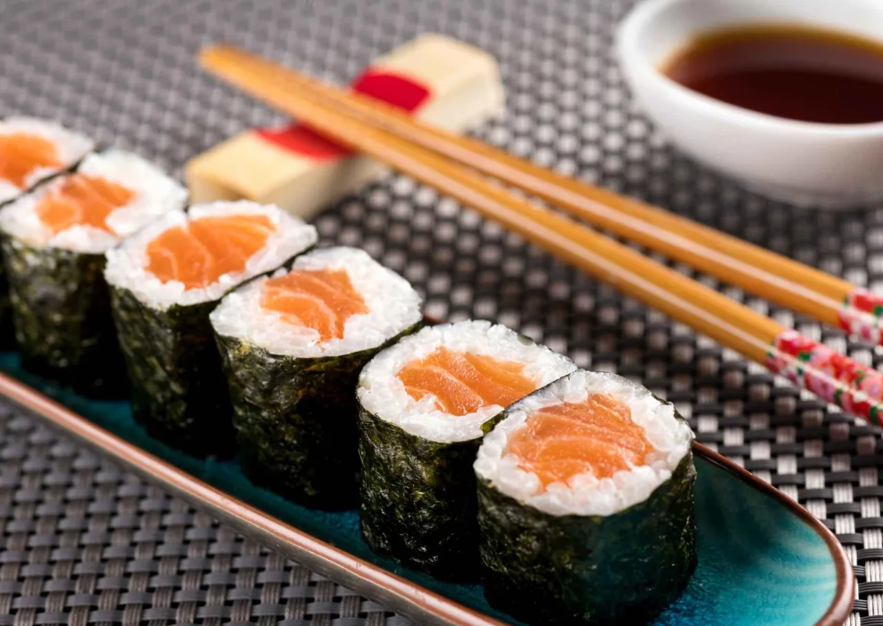 Rząd sushi z łososiem: kilka hosomaki i futomaki na talerzu, pałeczki i sos sojowy w tle.