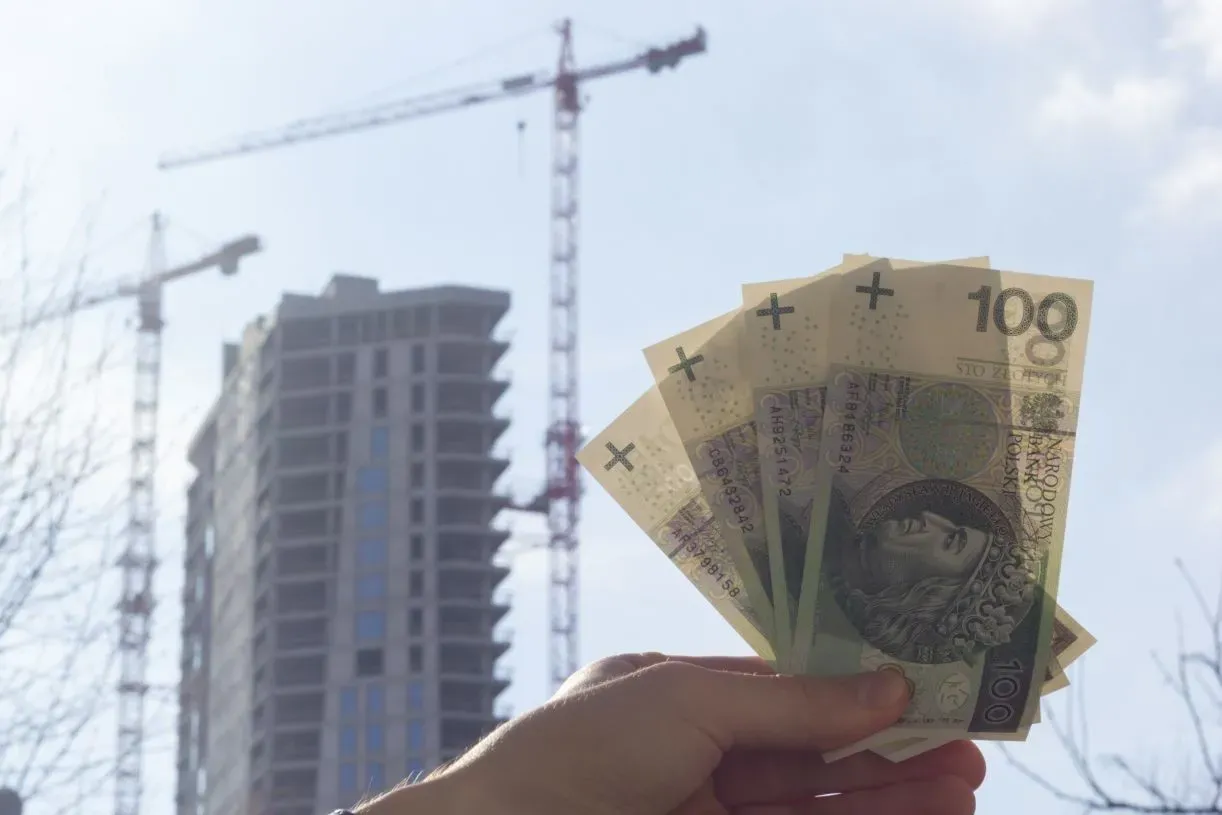 Dłoń trzyma banknoty 100 zł na tle budowy. Czy to symbol wsparcia dla programów rządowych mieszkaniowych?