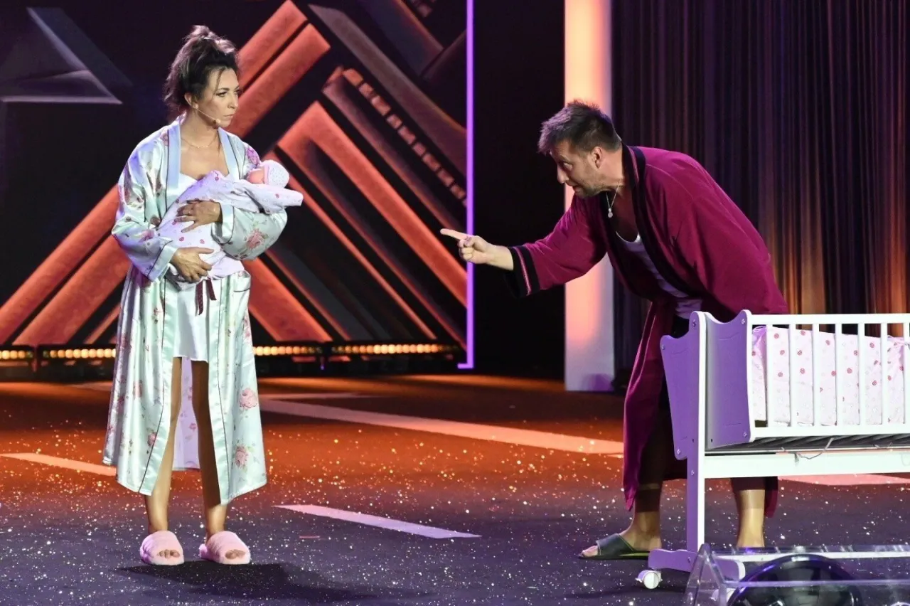 Scena z kabaretu z Kielc: kobieta w szlafroku tuli dziecko, mężczyzna w szlafroku wskazuje palcem na łóżeczko.