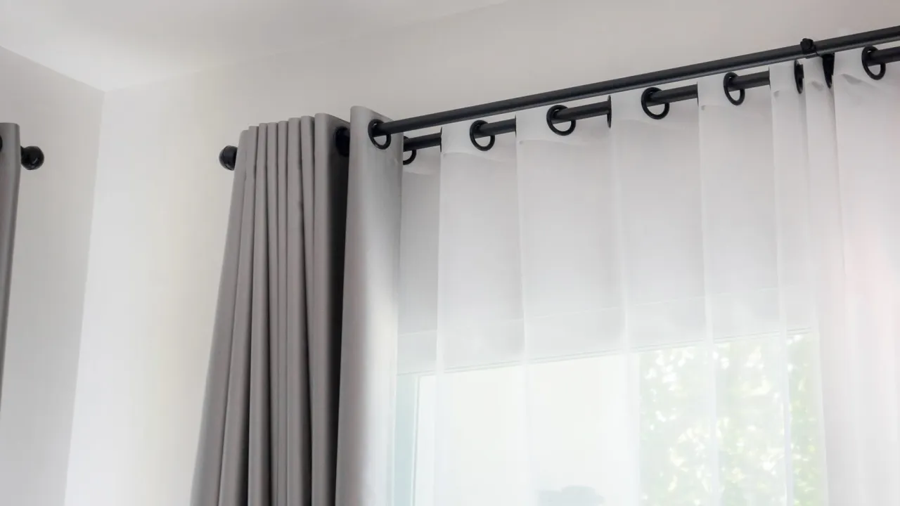 Szare i białe zasłony wiszą na czarnym karniszu, zamontowanym blisko sufitu. Okno jest zasłonięte przezroczystą tkaniną.