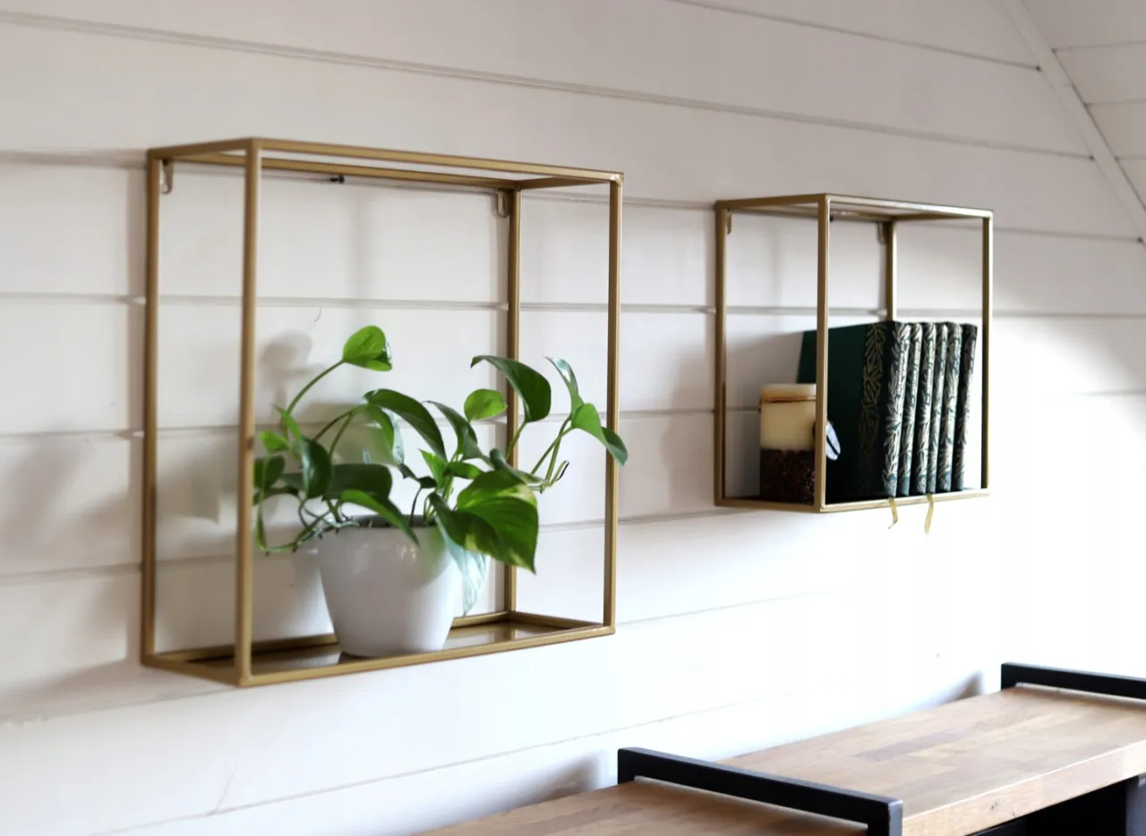 Złote ramki na ścianie, idealne, by stworzyć własny, stylowy kwietnik na ścianę. Jedna z nich zdobi zielona roślina, druga książki i świeca.