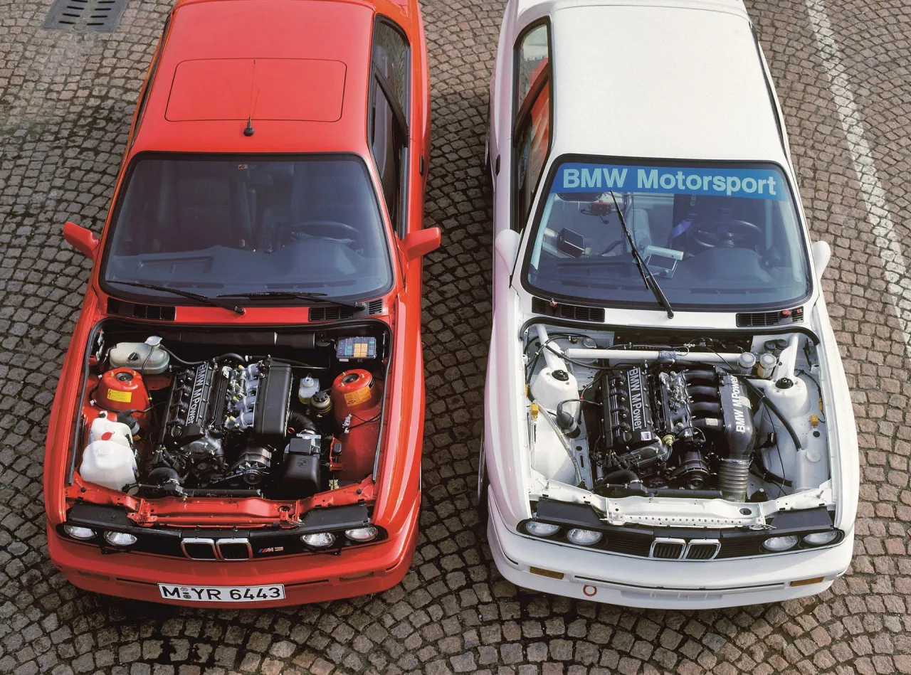 Dwa klasyczne BMW z otwartymi maskami, prezentujące moc silnik&oacute;w BMW M Power. Czerwony i biały model stoją obok siebie na brukowanej nawierzchni.