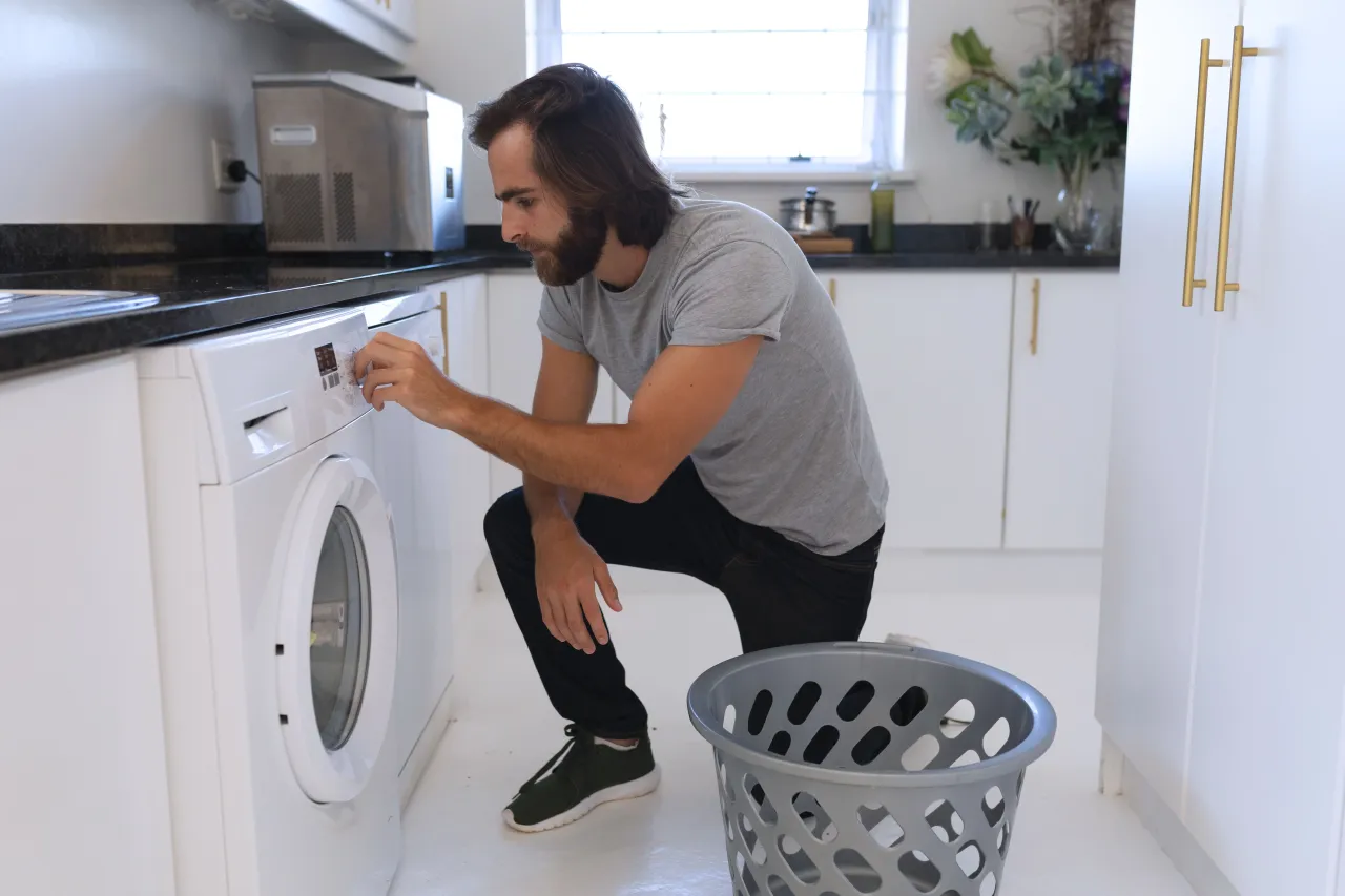 Mężczyzna przy pralce, przygotowujący się do prania, być może szukający sposobu, jak uratować skurczony sweter z wełny. Obok stoi kosz na pranie.