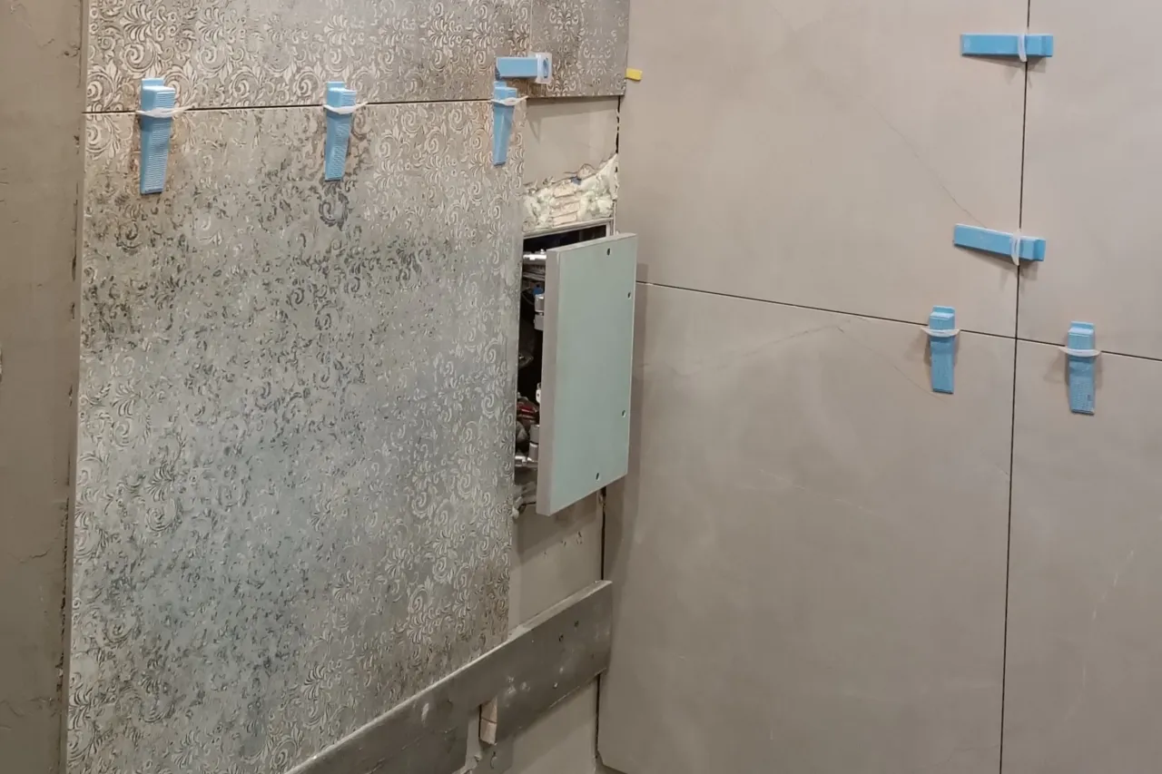 Montaż drzwiczek rewizyjnych plastikowych w łazience. Widoczne płytki, kliny do poziomowania i otwarta wnęka.