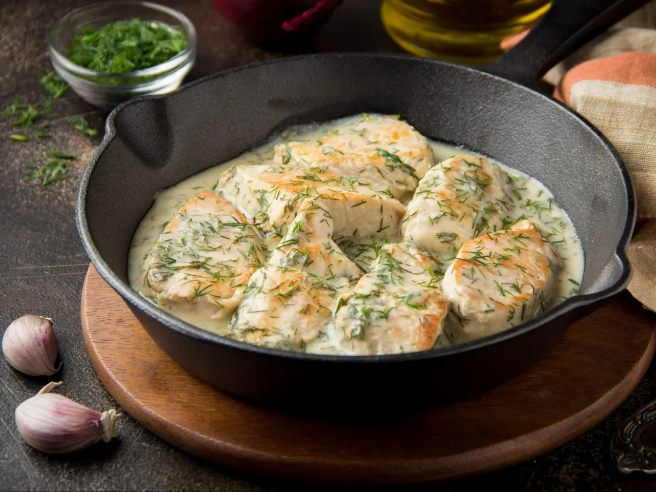 Polędwiczki drobiowe w kremowym sosie z koperkiem, podane w żeliwnej patelni. Obok czosnek i miseczka z posiekanym koperkiem.