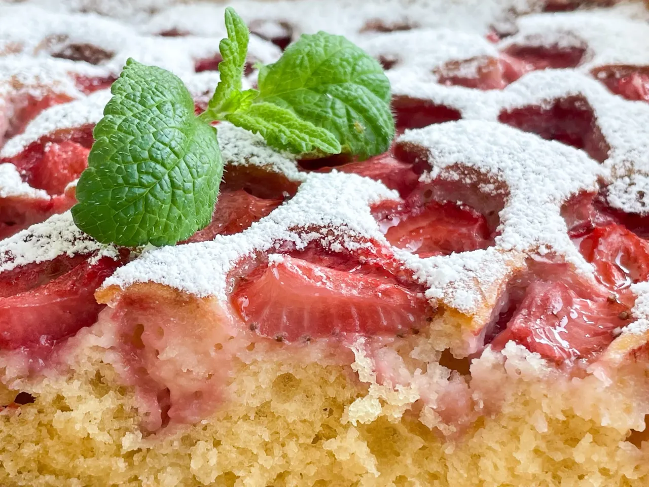 Puszyste ciasto jogurtowe z truskawkami, posypane cukrem pudrem i ozdobione listkami mięty. Idealne na letnie popołudnie.