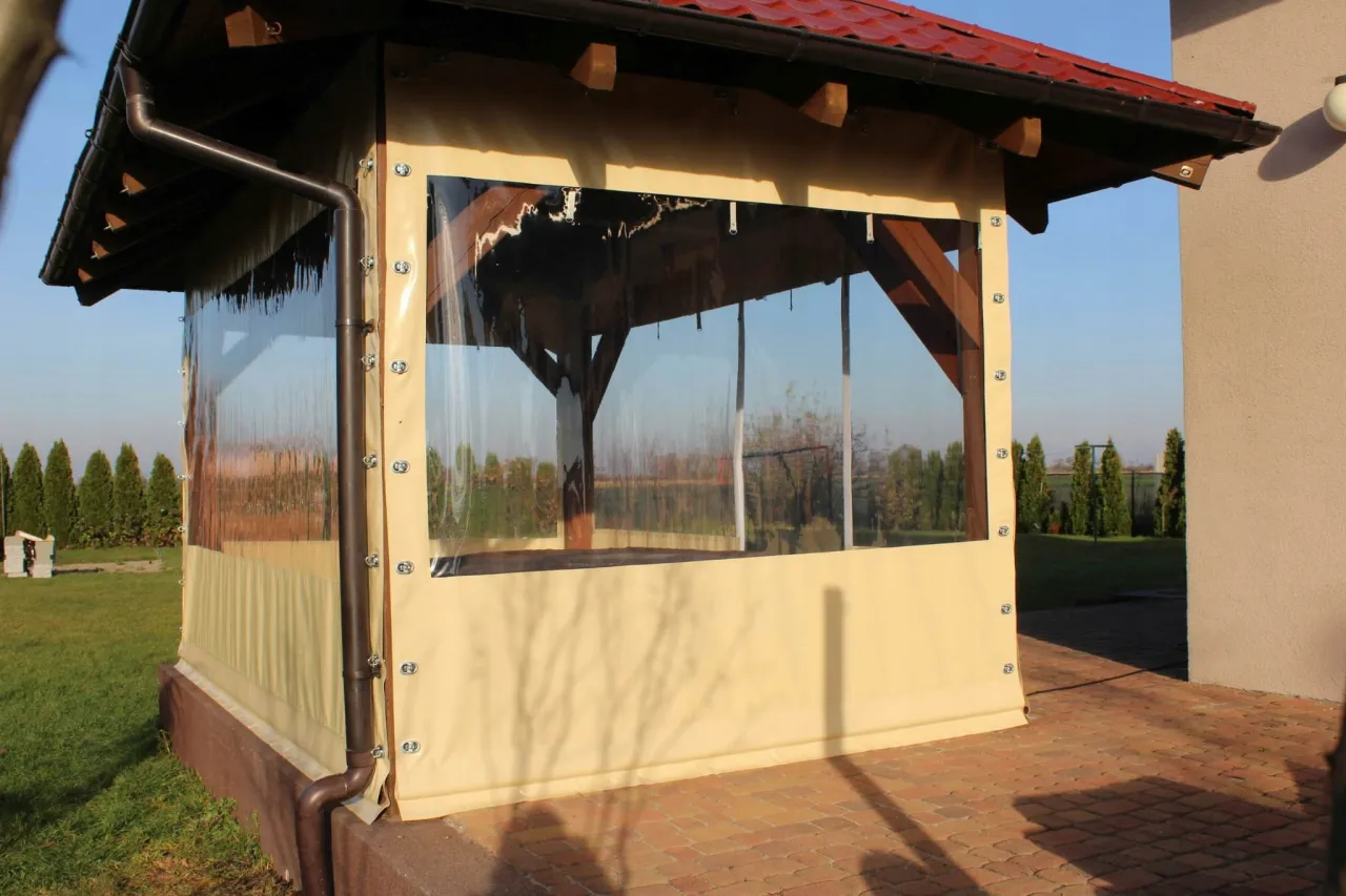 Zabezpieczona altanka z przezroczystymi zasłonami i beżowym materiałem u dołu, chroniąca przed wiatrem.